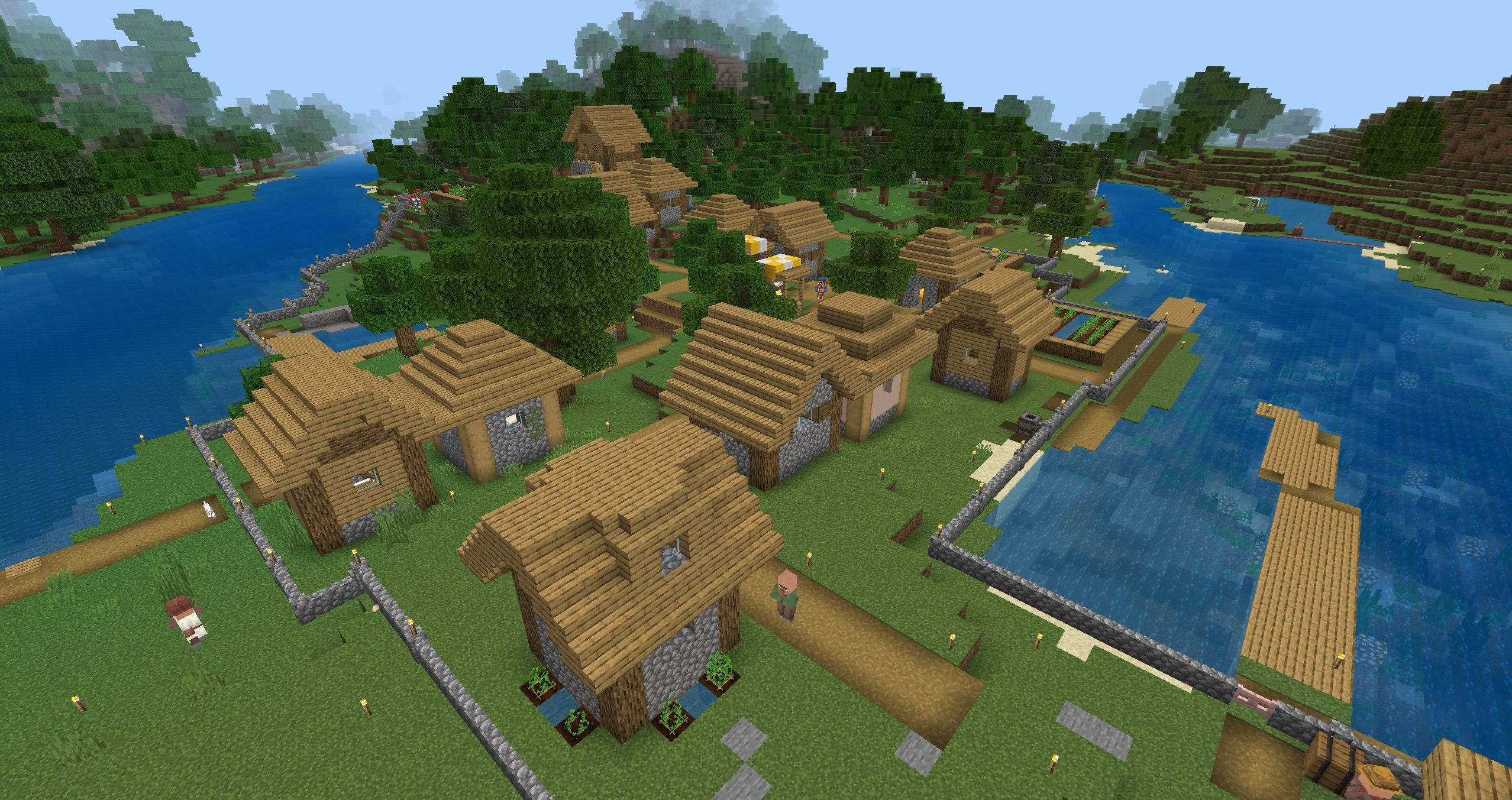 A view of a Village from overhead. It's dotted with trees, walled off, and surrounded by water on two sides in Minecraft.